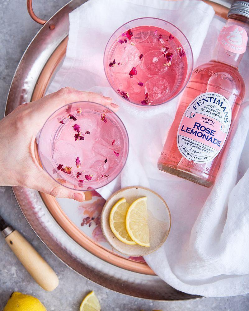 Pink rose lemonade in glasses with ice and petals, lemon slices, Fentimans bottle on tray