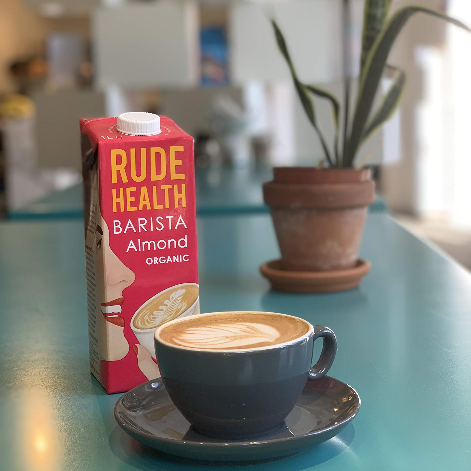Carton of Rude Health Barista Almond Organic milk next to a cup of coffee on a table.