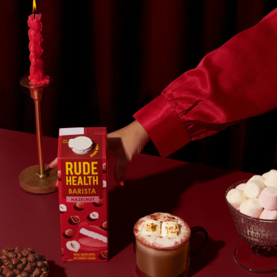 Rude Health Barista Hazelnut milk carton on a red table with hot chocolate and marshmallows.