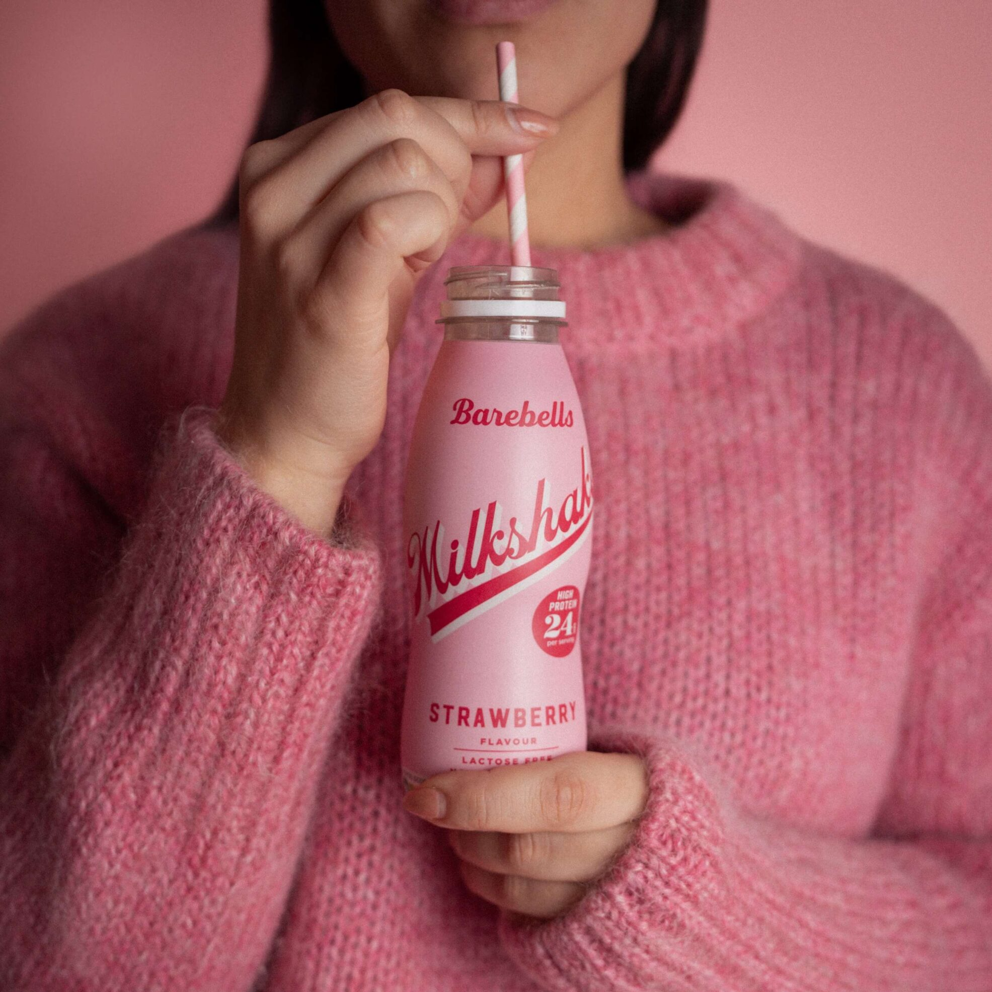 Person holding a Barebells Strawberry Milkshake bottle with a pink background