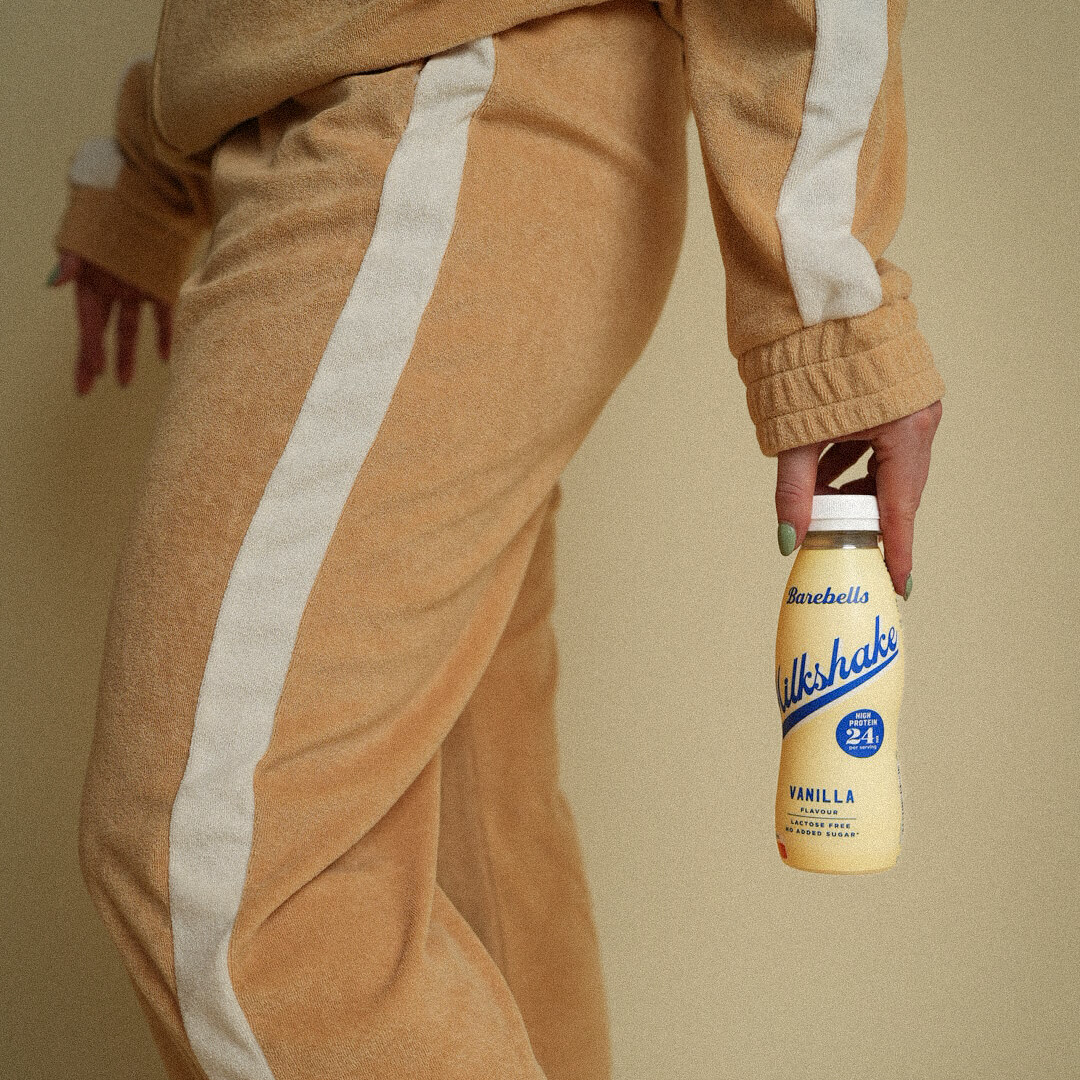 Person wearing a brown tracksuit with white stripes holding a bottle of Milkshakes vanilla ice cream.