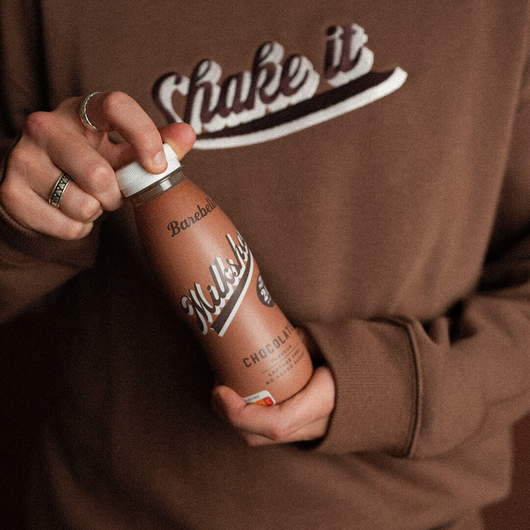 Person wearing a brown sweatshirt with 'Shake it' text, holding a chocolate milk bottle.
